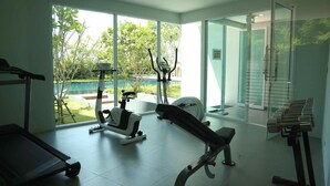 Un gimnasio con diversos equipos de ejercicio, ventanas amplias y vistas a una piscina y zonas verdes.
