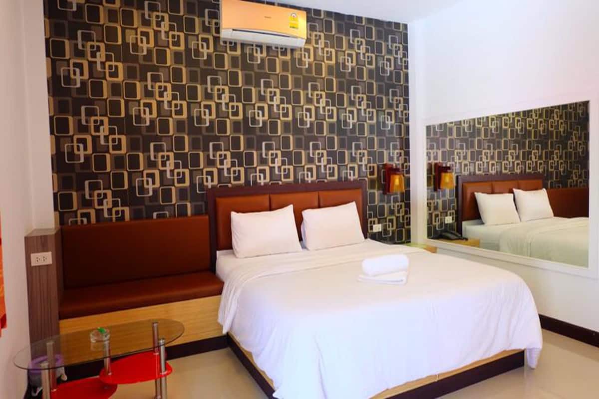 A hotel room with a patterned wall, a bed with white linens, a brown sofa, and a glass table.