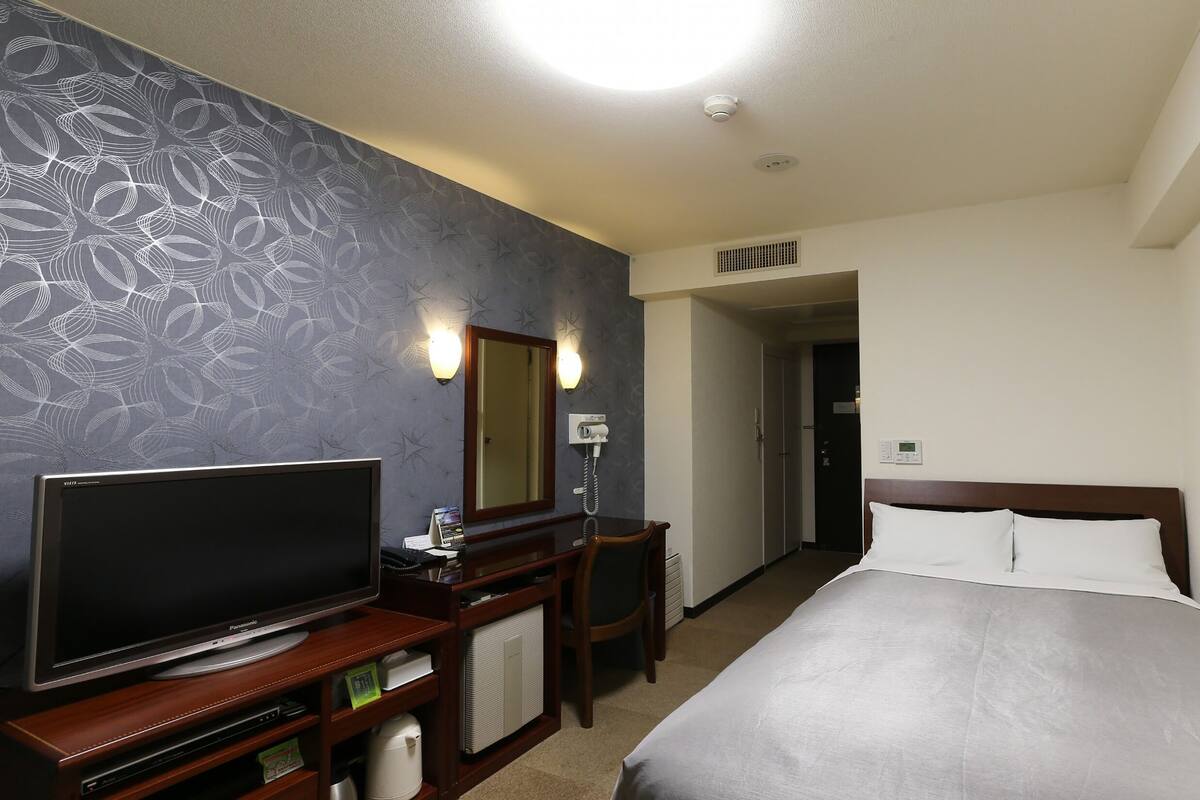 Habitación de hotel con una amplia televisión de pantalla plana, un escritorio con lámpara, un espejo, una cama con almohadas blancas y un papel tapiz estampado.
