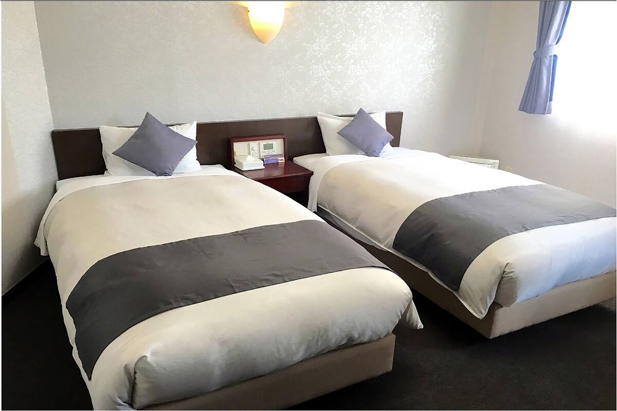 Habitación de hotel con dos camas, una mesita de noche, un reloj de pared y una ventana con cortinas.