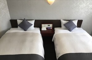 Habitación de hotel con dos camas, cada una con ropa blanca y almohadas oscuras. Hay una mesita de noche entre las camas con una lámpara y una pequeña mesa con un teléfono y una caja.