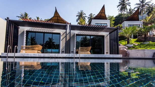 Un espace moderne aménagé au bord de la piscine, avec une piscine à eau claire, deux chaises longues et un bâtiment doté de grandes fenêtres et d’un toit recouvert de tuiles.