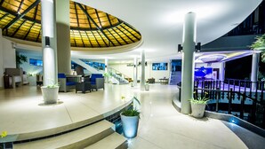 Un hall d’entrée spacieux avec un plafond haut, un escalier central et de nombreuses plantes en pot.
