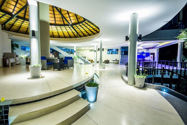 A spacious lobby with a high ceiling, a central staircase, and multiple potted plants.