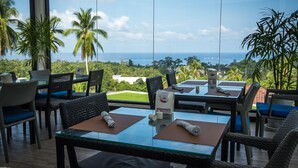 Un espace repas avec des tables et des chaises, une cloison en verre et une vue sur l’océan et les palmiers.