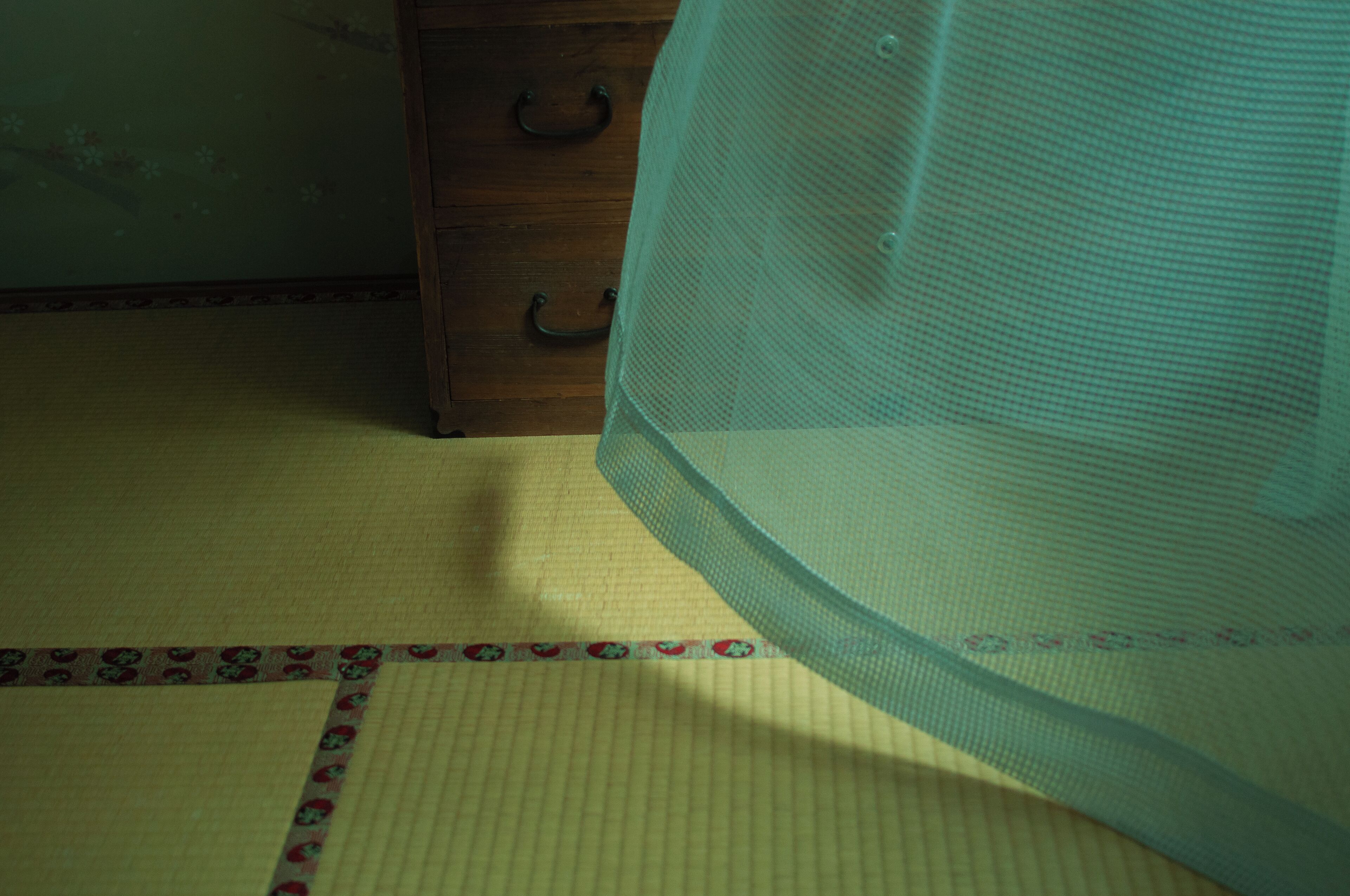 A room with tatami flooring, a wooden dresser with two drawers, and a mosquito net over a bed.