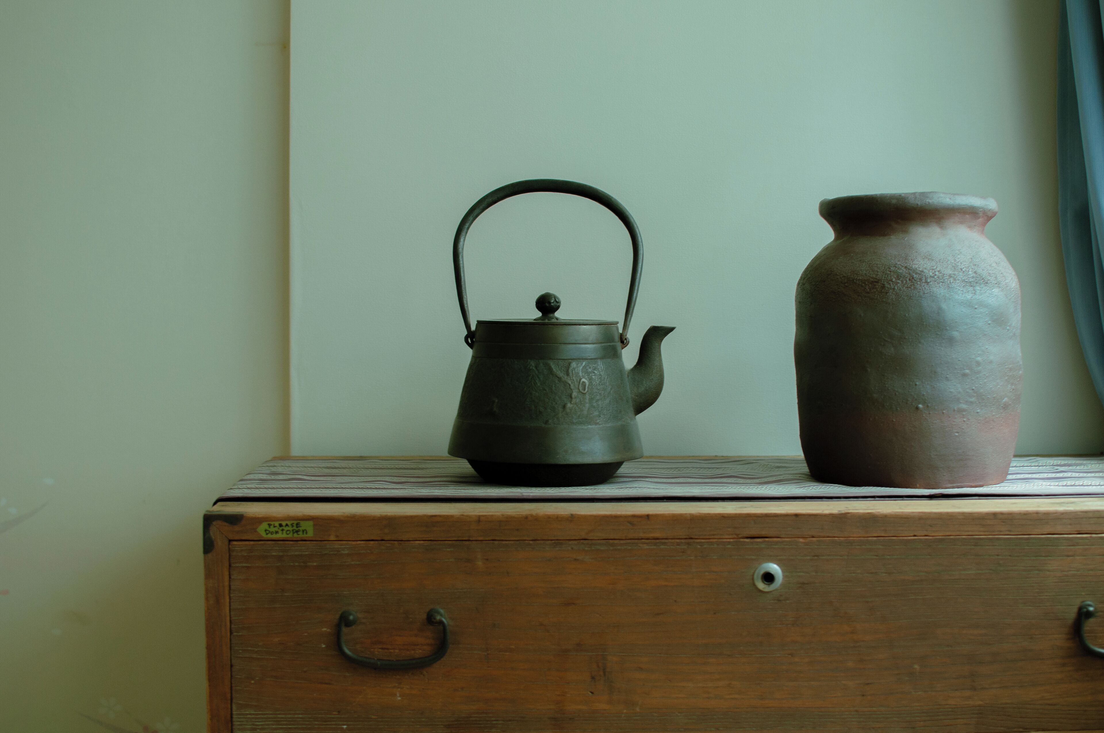 A wooden chest of drawers with a metal kettle and a clay pot on top.