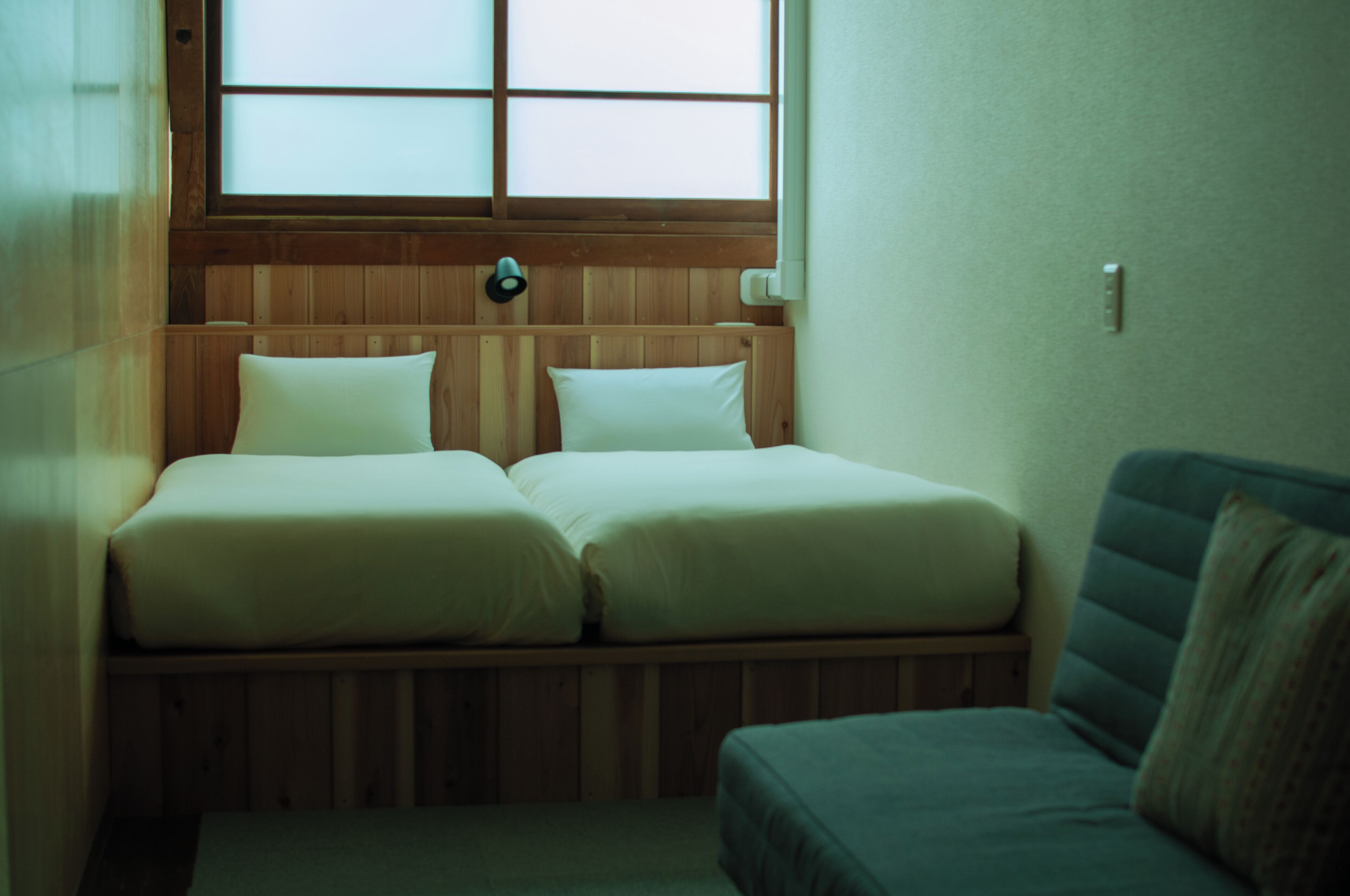 A small, compact room with two single beds, a wooden headboard, a window, and a green sofa.