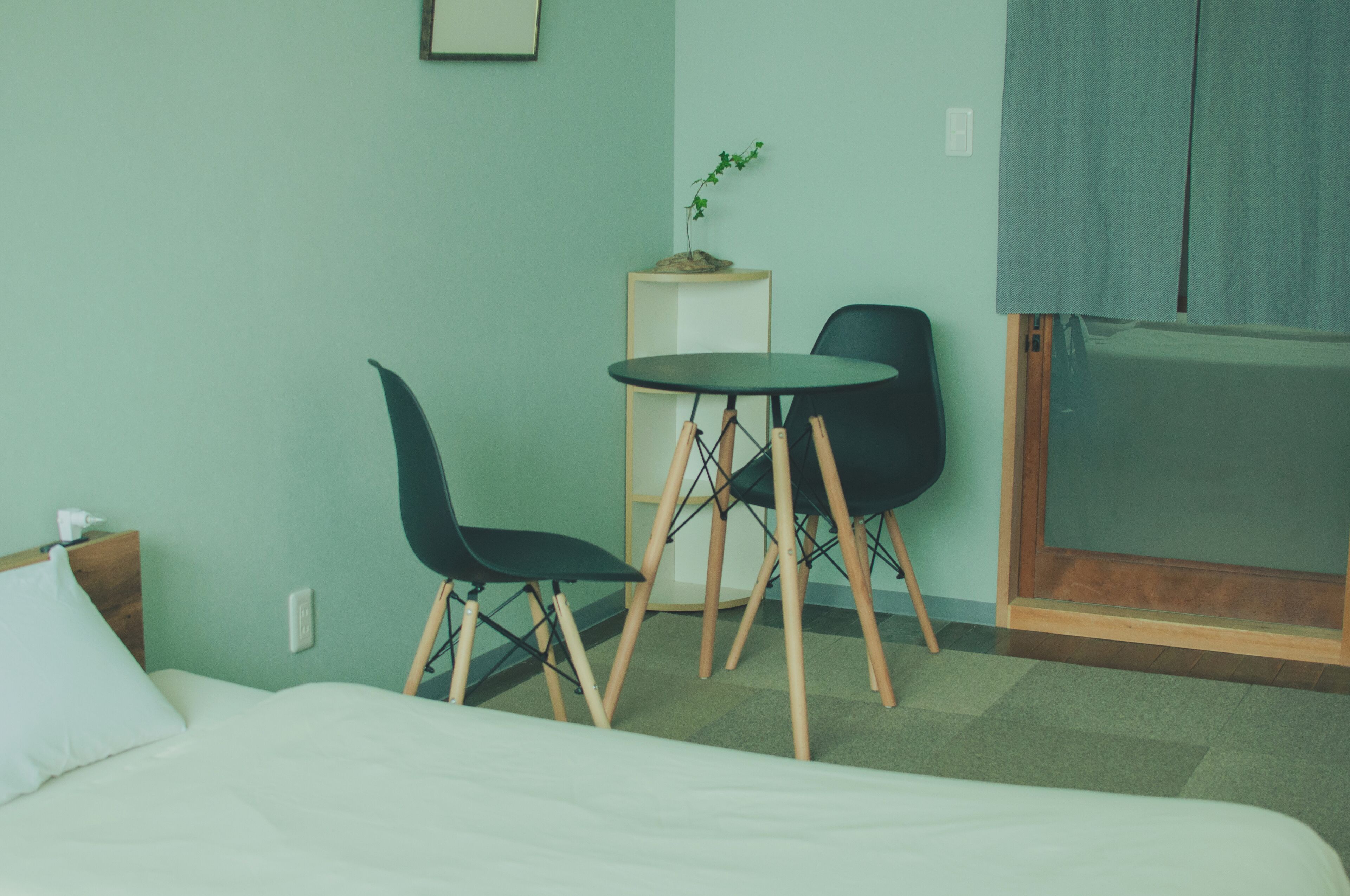 A hotel room with a bed, a small table with two chairs, a potted plant, and a view of the outside.