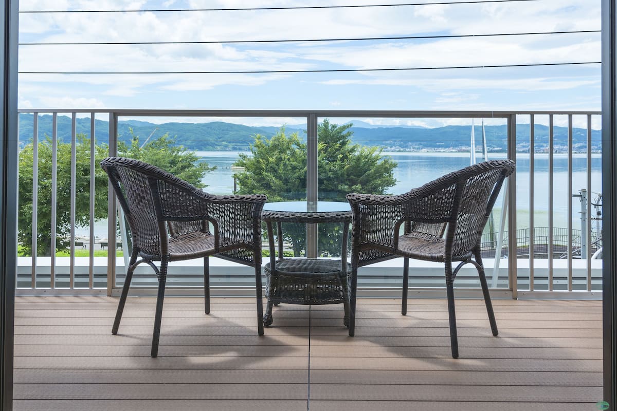 A balcony with wicker chairs and a small table, overlooking a lake and distant mountains.