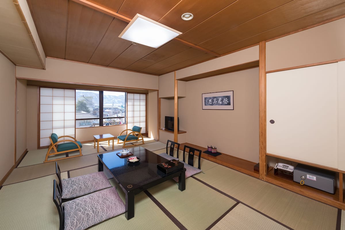 A traditional Japanese room with tatami flooring, a low table with cushions, a television, and a sliding door with a view of mountains.