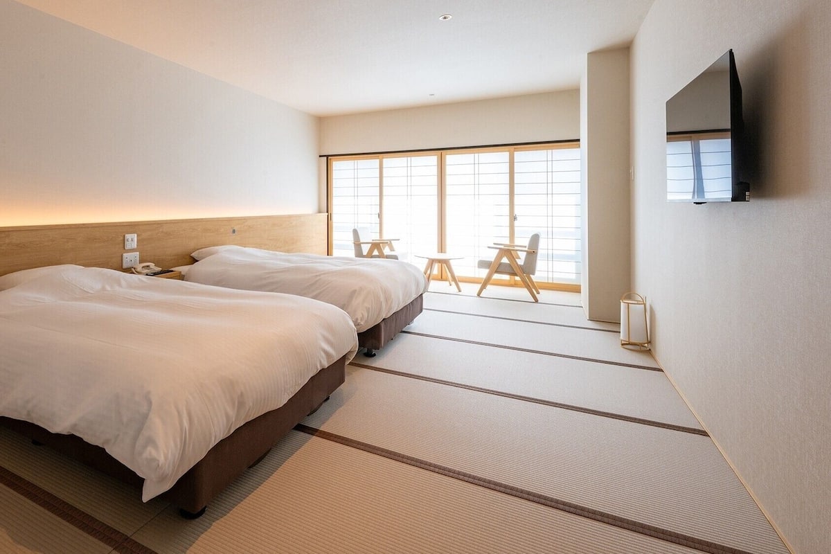 A hotel room with two beds, a TV, a window with a view, and a small table with chairs.