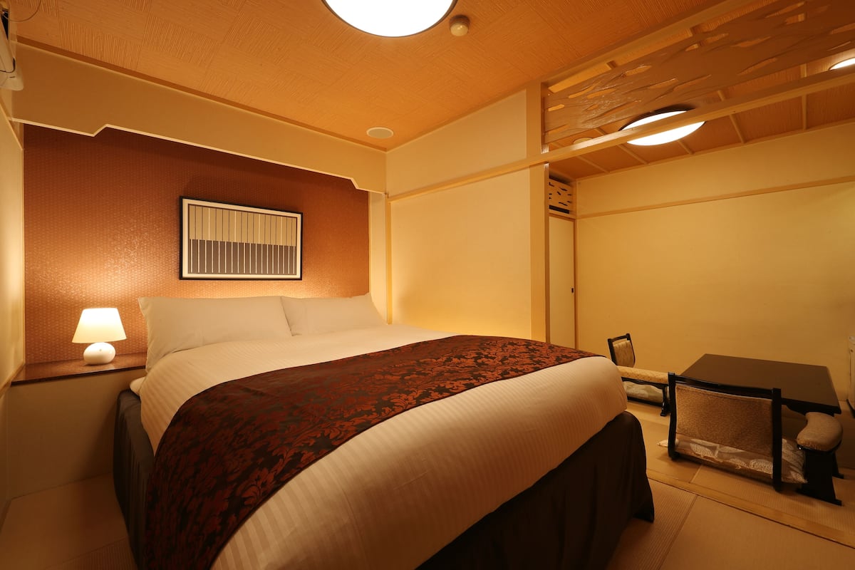 Quarto de hotel com uma cama, mesa de cabeceira com luminária, uma mesinha e uma cadeira.