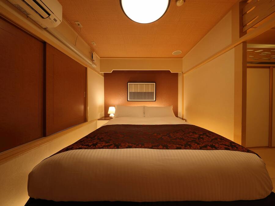 Ein Hotelzimmer mit einem großen Bett, einem Nachttisch mit Lampe, einem Fenster mit Jalousien und einem Deckenventilator.