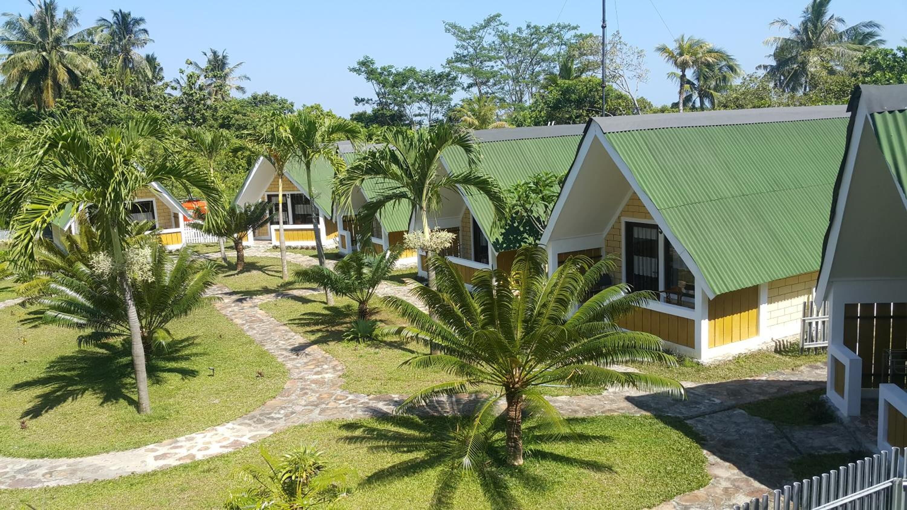 A row of bungalow-style cottages with green roofs, surrounded by palm trees and a stone pathway.