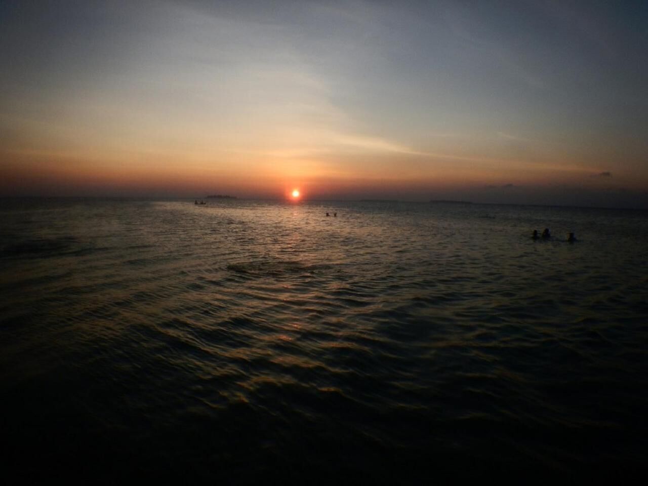 A sunset over a calm sea with a few people in the water.