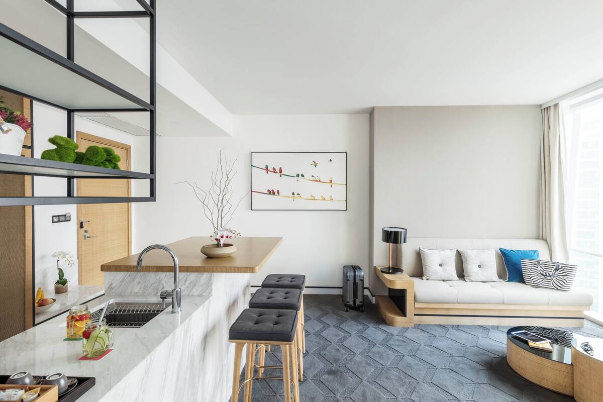 Une cuisine moderne avec un plan de travail en marbre, un évier et une étagère intégrée. On y trouve un coin salon avec un canapé et une table basse. Un tableau représentant des oiseaux est accroché au mur.
