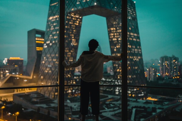 A person standing in front of a large window with a cityscape view, including a distinctive building with a circular pattern.