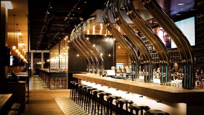 A bar with a long counter and multiple stools, a high ceiling with exposed pipes, and a patterned floor.