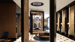 A modern lobby with a geometric patterned floor, a central seating area with a tree sculpture, and a large round clock on the ceiling.