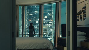 A bedroom with a large bed, a TV, and a view of a cityscape through floor-to-ceiling windows.