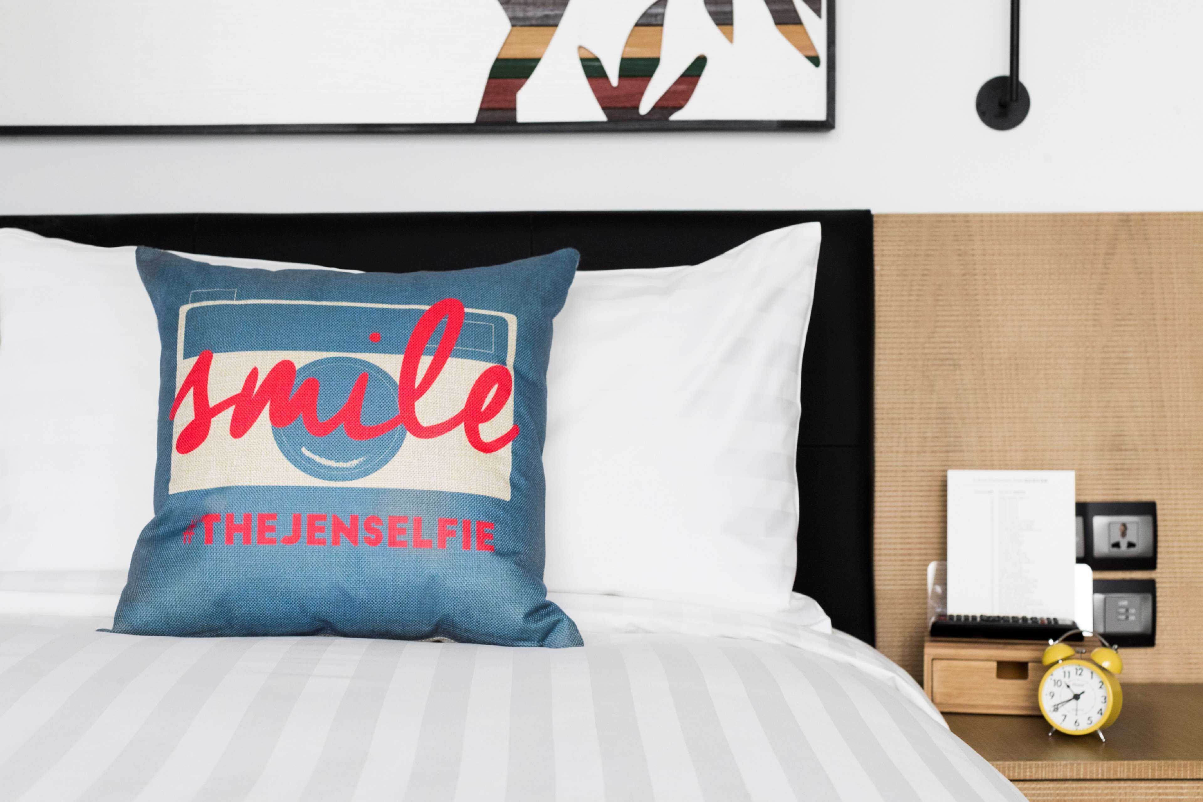 A neatly made bed with a pillow featuring a smiley face and the text "smile #THEJENSELFIE".
