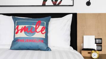 A neatly made bed with a pillow featuring a smiley face and the text "smile #THEJENSELFIE".