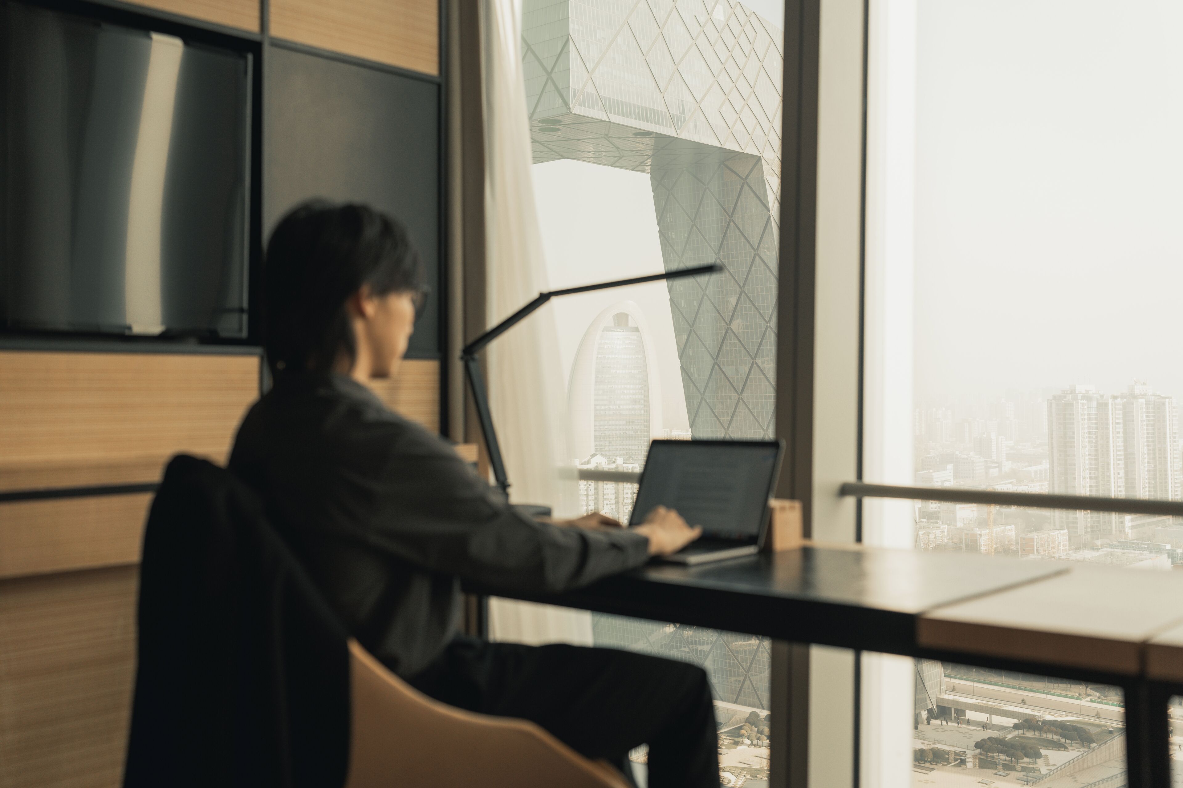 A person working at a desk with a laptop, large windows, and a view of a cityscape.