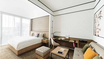 A modern hotel room with a large bed, a desk, a sofa, and a window with a city view.