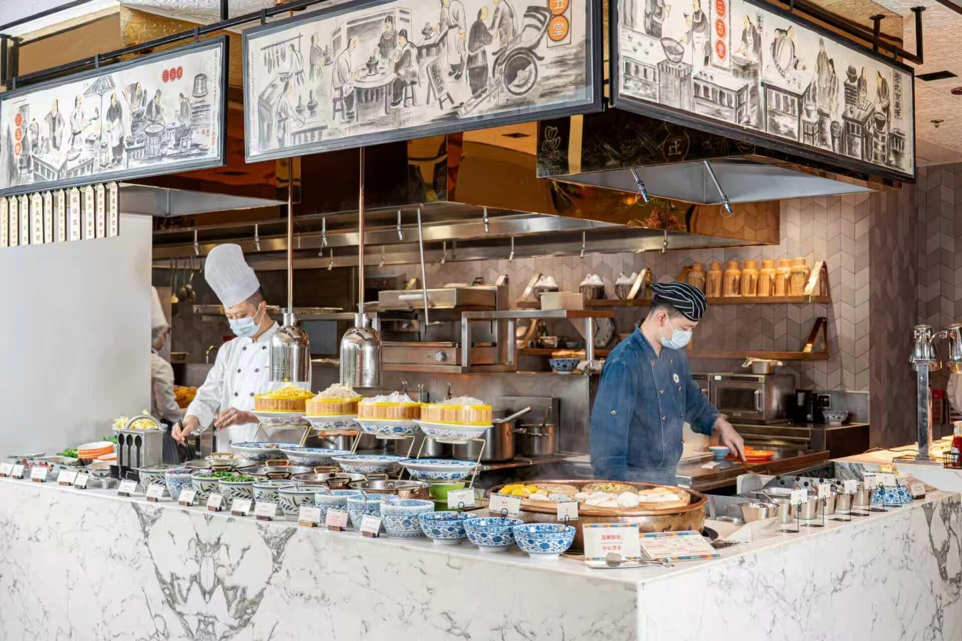 A restaurant with chefs preparing food, a marble counter with dishes, and traditional artwork on the walls.