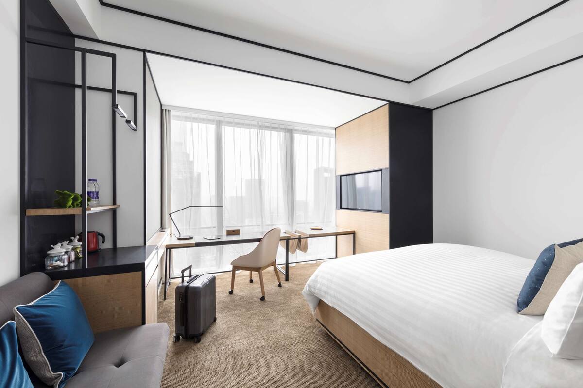 Une chambre d’hôtel moderne dotée d’un grand lit, d’un bureau avec une chaise, d’un petit coin salon et d’une grande fenêtre donnant sur la ville.