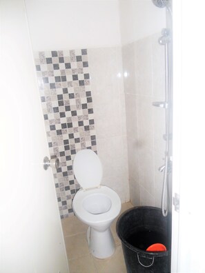A bathroom with a tiled wall, a toilet, a shower, and a black bucket. - Bromo Sunrise Homestay (Probolinggo)