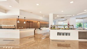 A modern hotel lobby with a central reception desk, a seating area with a view of the outdoors, and a buffet counter with various food items. - Globales Cala Bona (Son Servera)