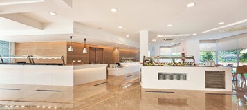 Un moderno vestíbulo de hotel con un mostrador de recepción central, una zona de estar con vistas al exterior y una mesa de buffet con diversas opciones de comida.