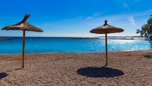 Dois guarda-sóis de palha numa praia de seixos com mar azul límpido e afloramentos rochosos ao longe.