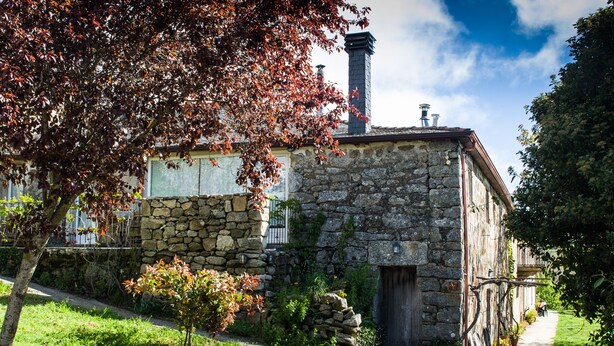 Un edificio de piedra con chimenea, un camino y un árbol con hojas rojas.