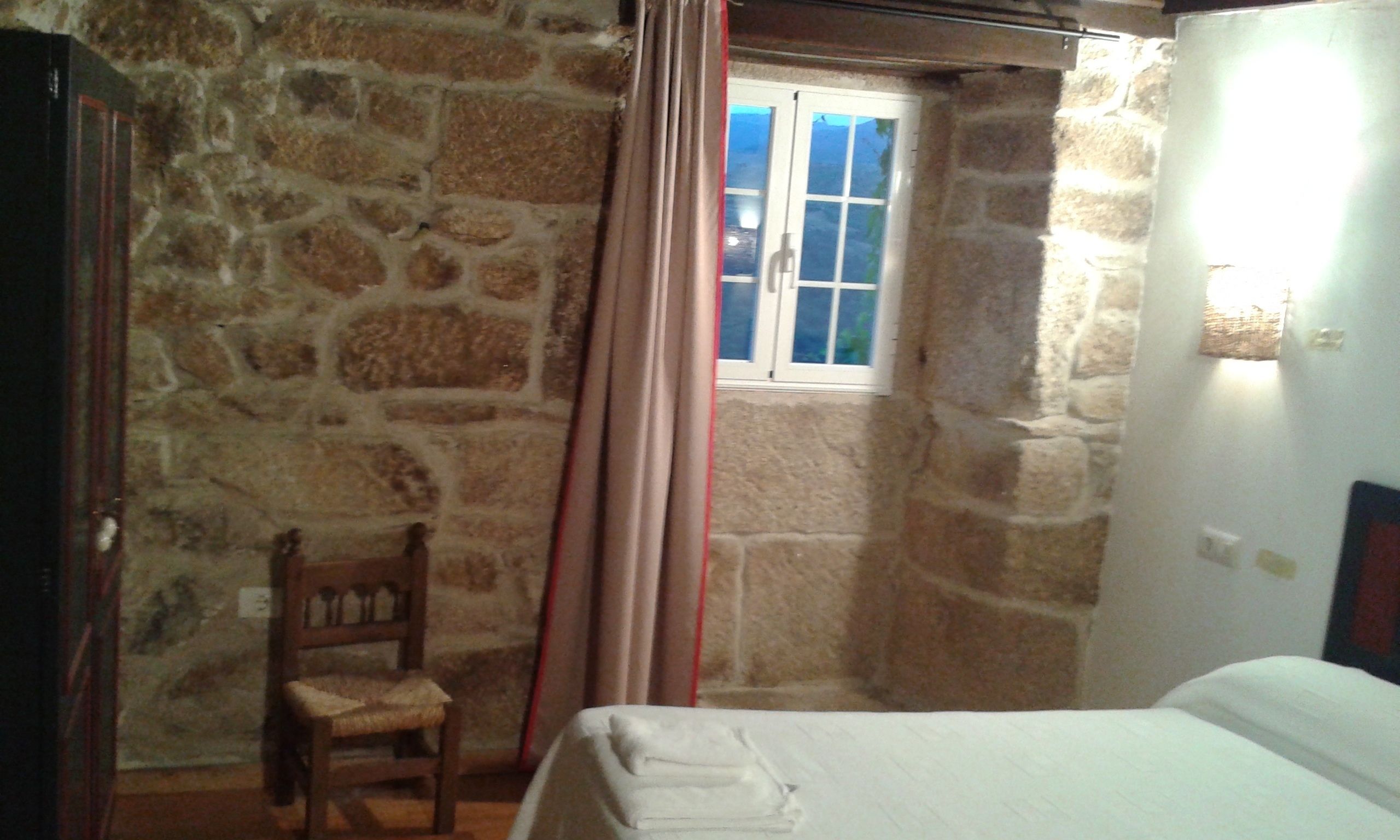 A stone-walled room with a bed, a chair, and a window with curtains.
