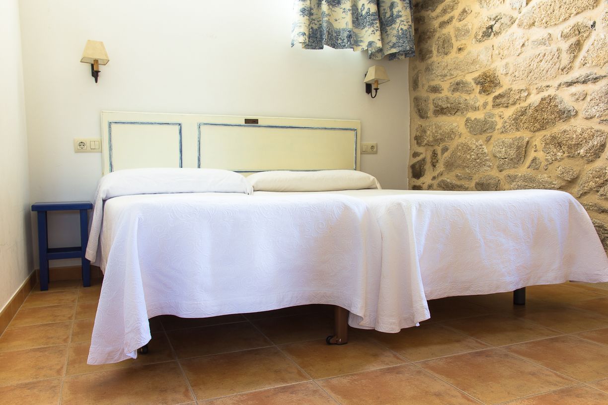 Un lit simple avec une couvre-lit blanche, dans une pièce dont un mur est en pierre et qui contient un petit tabouret bleu.