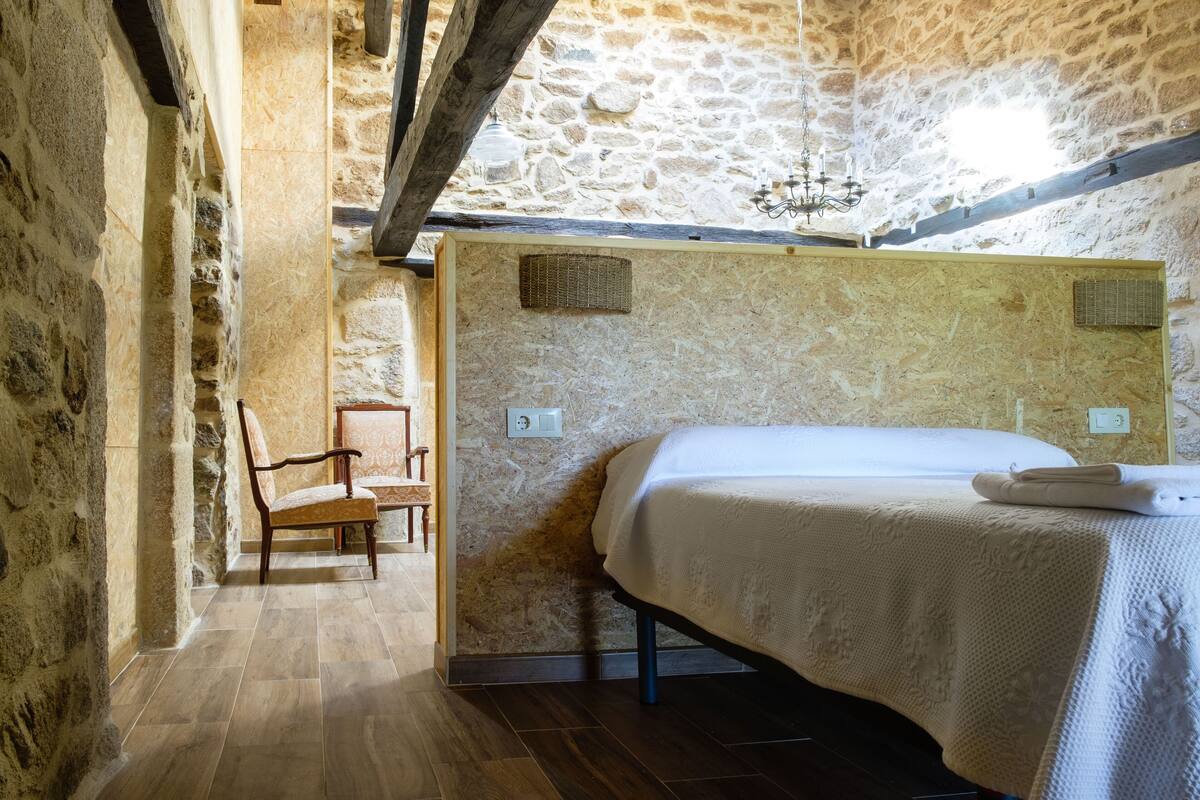 A bedroom with stone walls, a wooden bed frame, a chair, and a chandelier.