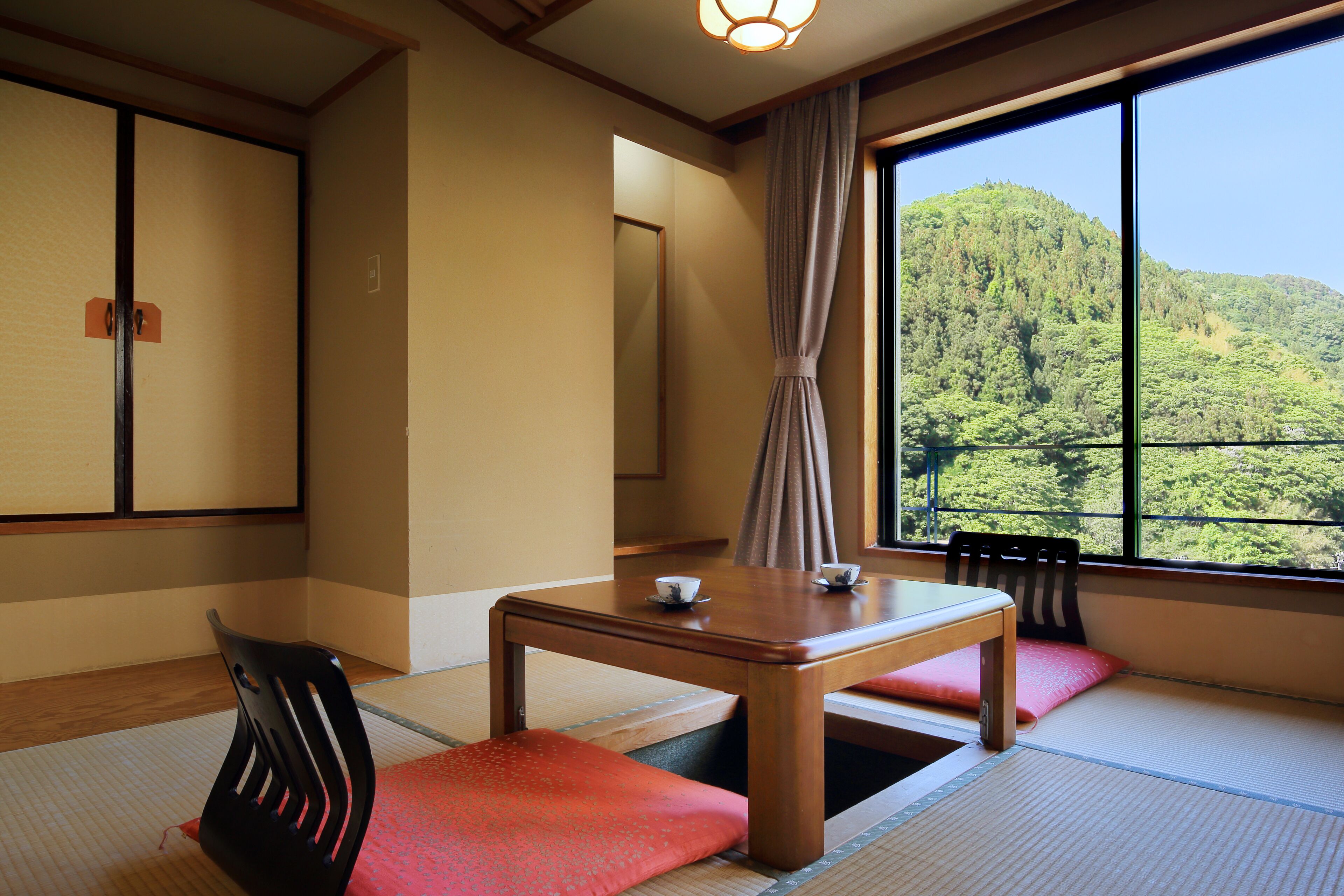 A traditional Japanese room with tatami flooring, a low table with two cups, a chair, and a view of a forested hill through a large window.