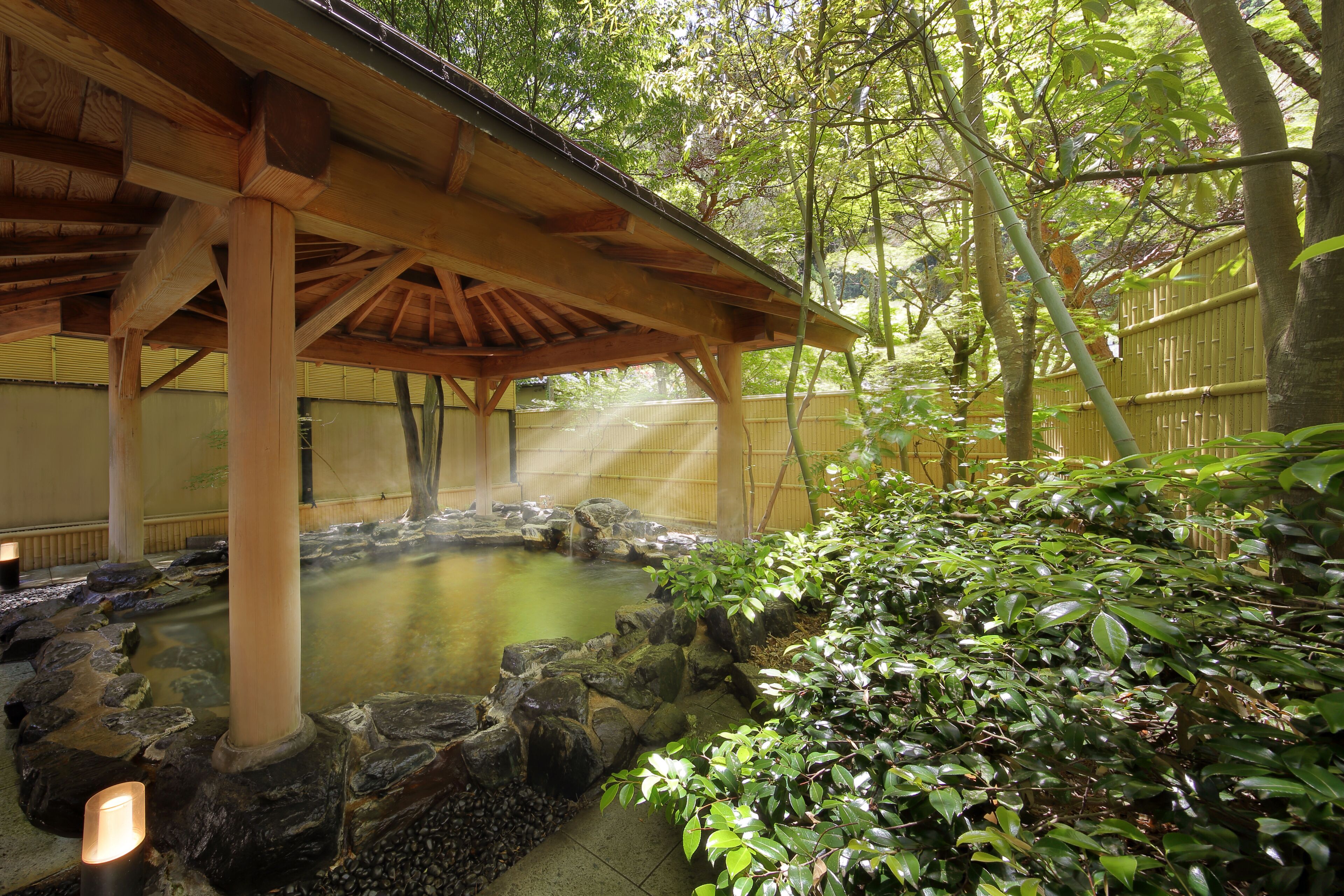Un onsen japonés tradicional con un pabellón de madera, un manantial termal rodeado de rocas y abundante vegetación.
