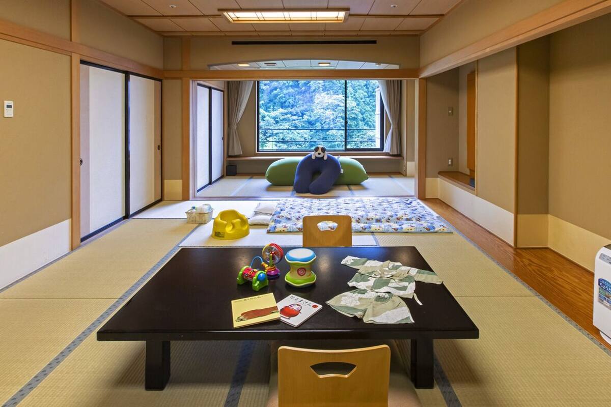 Ett traditionellt japanskt rum med tatamimattor på golvet, en futonmadrass, ett fönster med utsikt över träd, ett bord med barnleksaker och en värmare.