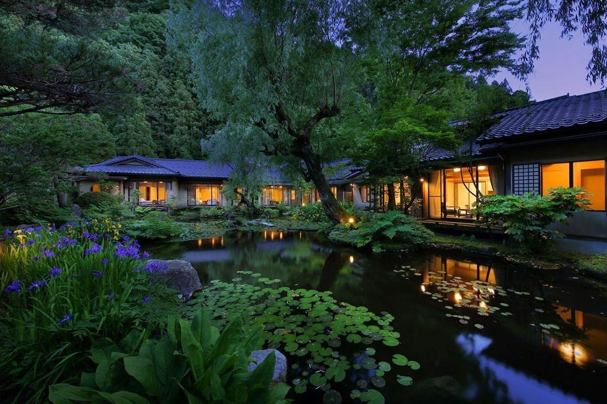 Un edificio de estilo japonés tradicional con un estanque, rodeado de exuberante vegetación e iluminado con una cálida luz.