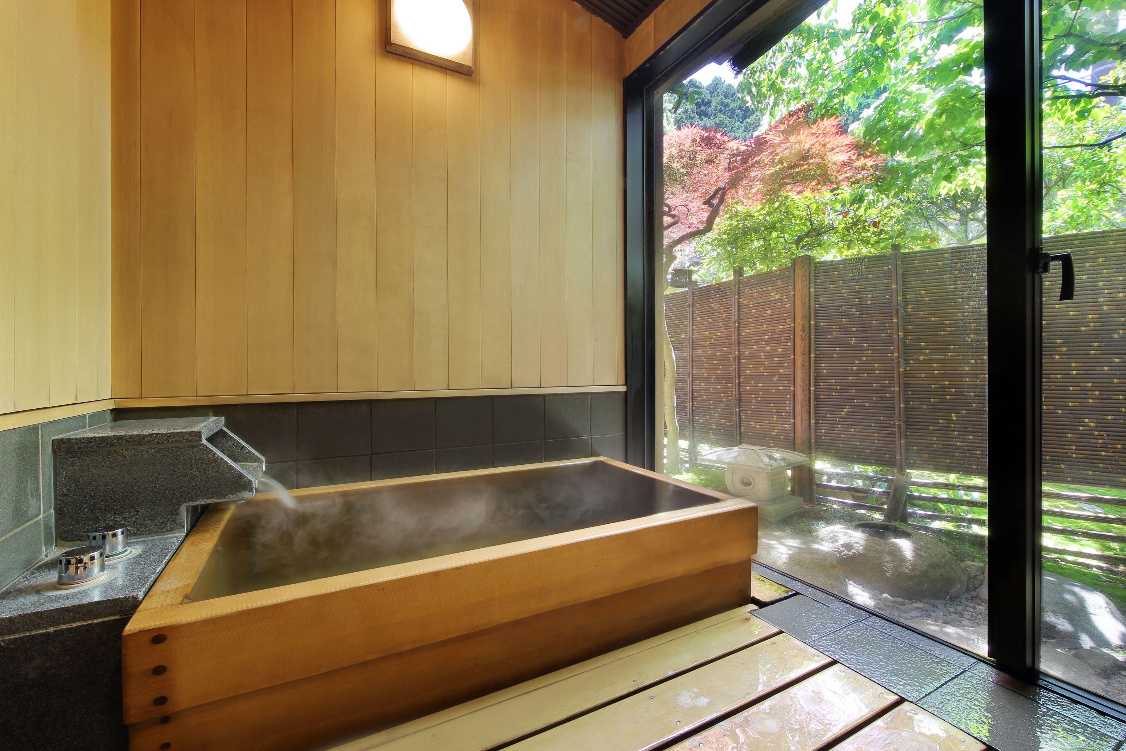 A wooden bathtub with steam, a tiled wall, and a view of a garden with trees.