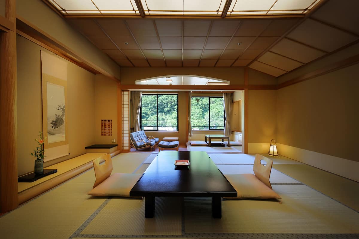 Ett traditionellt japanskt rum med tatamimattor på golvet, ett lågt bord, kuddar och utsikt över grönskan genom stora fönster.