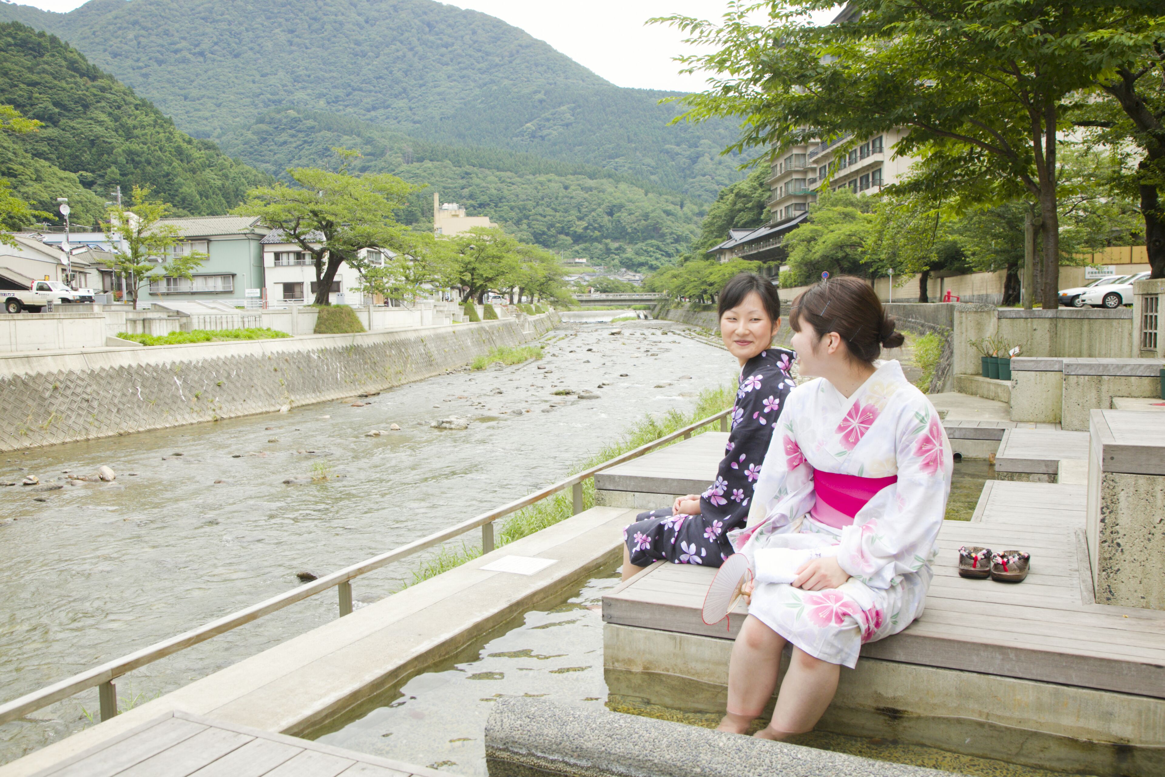 Two individuals seated on a wooden platform by a river, with buildings and greenery in the background.