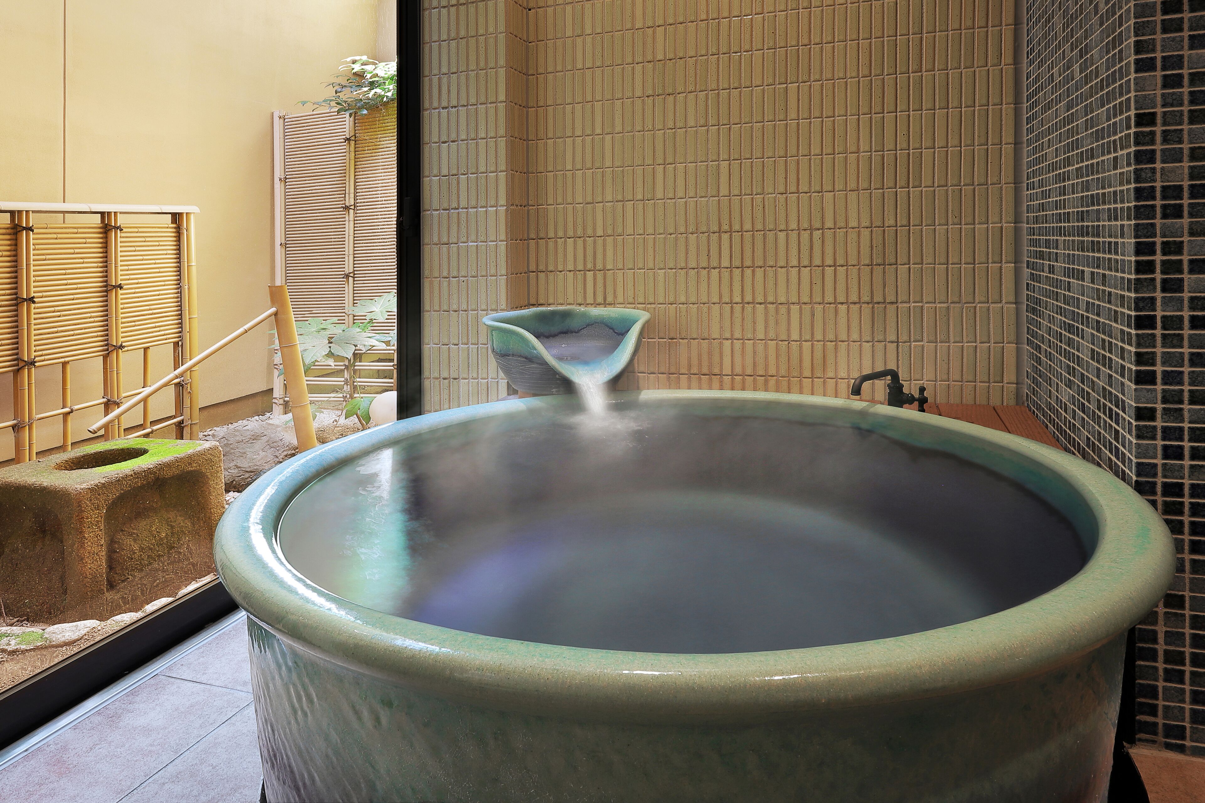 Un grand bain à remous en céramique, surmonté d’un petit récipient décoratif, entouré de bambous et bordé de murs recouverts de carreaux.