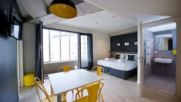 A modern hotel room with a bed, a dining table with chairs, a large window, and a bathroom with a sink.