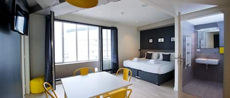 A modern hotel room with a bed, a dining table with chairs, a large window, and a bathroom with a sink.