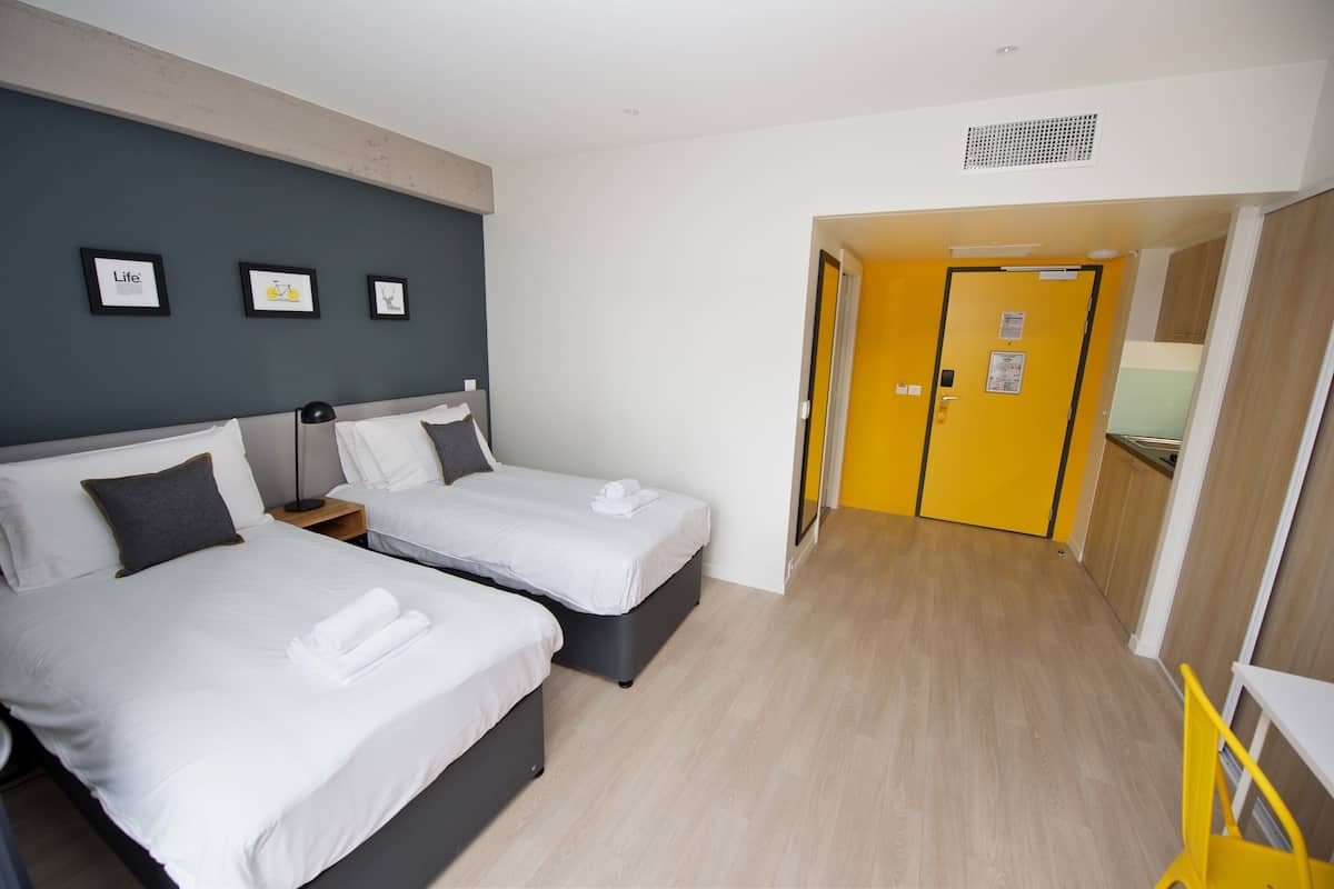 A hotel room with two beds, a small desk, and a yellow door leading to a bathroom.